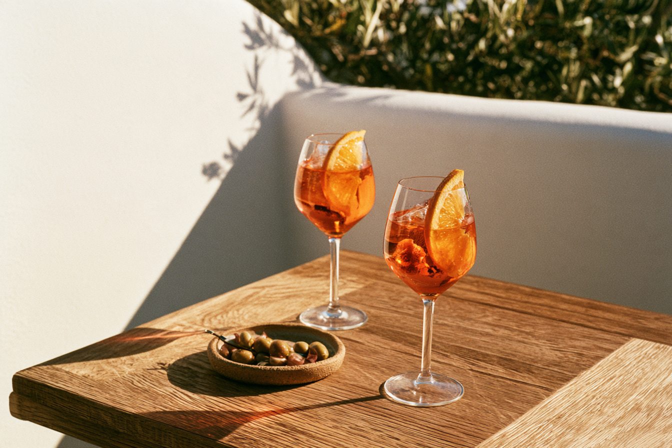 Two spritz cocktails and a bowl of olives on an oak table in low evening sun