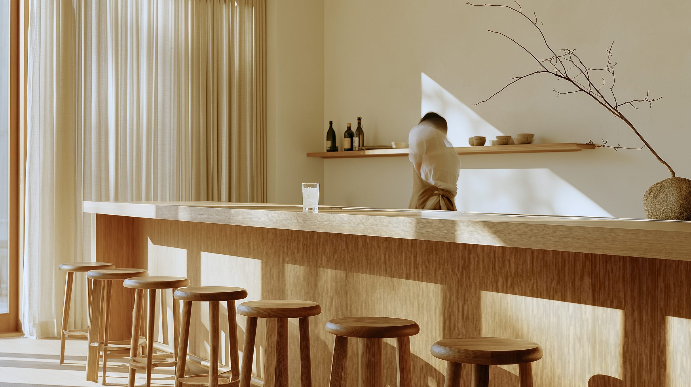 Sunlit oak bar counter with round wooden stools, a bartender reaching for a bottle on the shelf